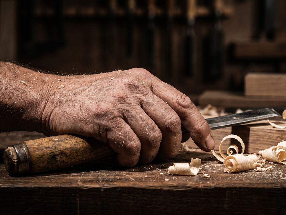 Main d'artisan burinée par le temps, posée sur l'établi avec un ciseau à bois, au milieu des copeaux